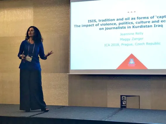 woman standing on stage speaking gesturing to a presentation behind her