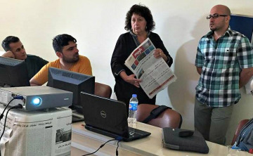 woman holding newspaper standing with three other people behind a table