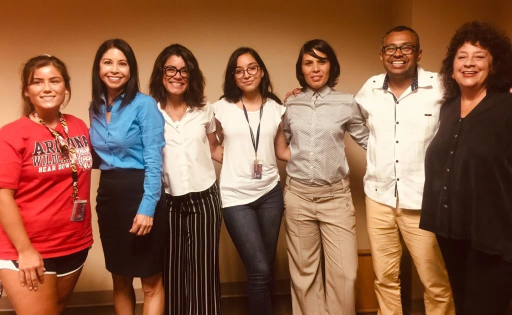seven people standing in a row with arms around each others' shoulders, indoors
